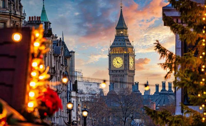 A dramatic, low-angle view of London's Big Ben standing centrally under a colourful sunset sky filled with pink, orange, and blue clouds. The clock face is warmly illuminated. In the foreground, out-of-focus Christmas tree branches decorated with glowing golden fairy lights frame both the left and right sides of the image, adding a warm holiday atmosphere to the urban cityscape.