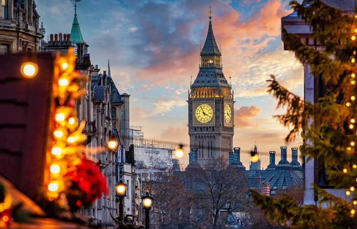 A dramatic, low-angle view of London's Big Ben standing centrally under a colourful sunset sky filled with pink, orange, and blue clouds. The clock face is warmly illuminated. In the foreground, out-of-focus Christmas tree branches decorated with glowing golden fairy lights frame both the left and right sides of the image, adding a warm holiday atmosphere to the urban cityscape.