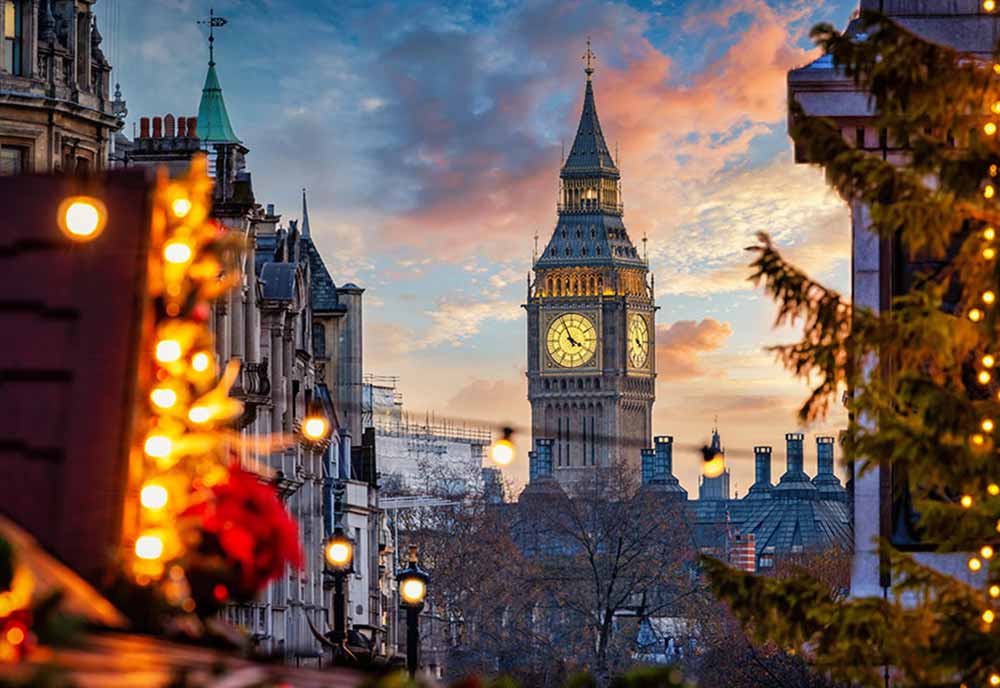 A dramatic, low-angle view of London's Big Ben standing centrally under a colourful sunset sky filled with pink, orange, and blue clouds. The clock face is warmly illuminated. In the foreground, out-of-focus Christmas tree branches decorated with glowing golden fairy lights frame both the left and right sides of the image, adding a warm holiday atmosphere to the urban cityscape.