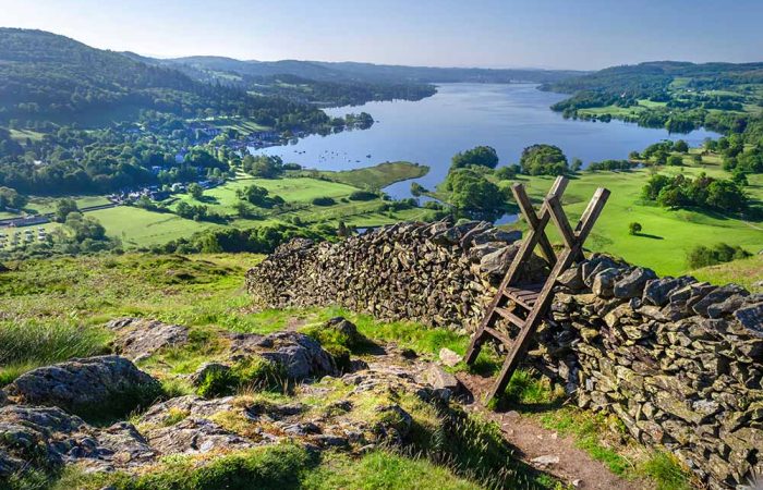 Elevated viewpoint of a dry stone wall with a wooden stile on a grassy hillside, overlooking a large lake surrounded by rolling green hills and countryside.