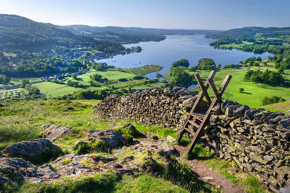 Elevated viewpoint of a dry stone wall with a wooden stile on a grassy hillside, overlooking a large lake surrounded by rolling green hills and countryside.